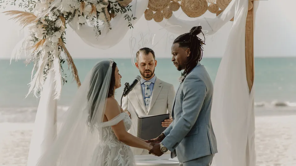 A couple holding hands during a beach wedding ceremony with an officiant.