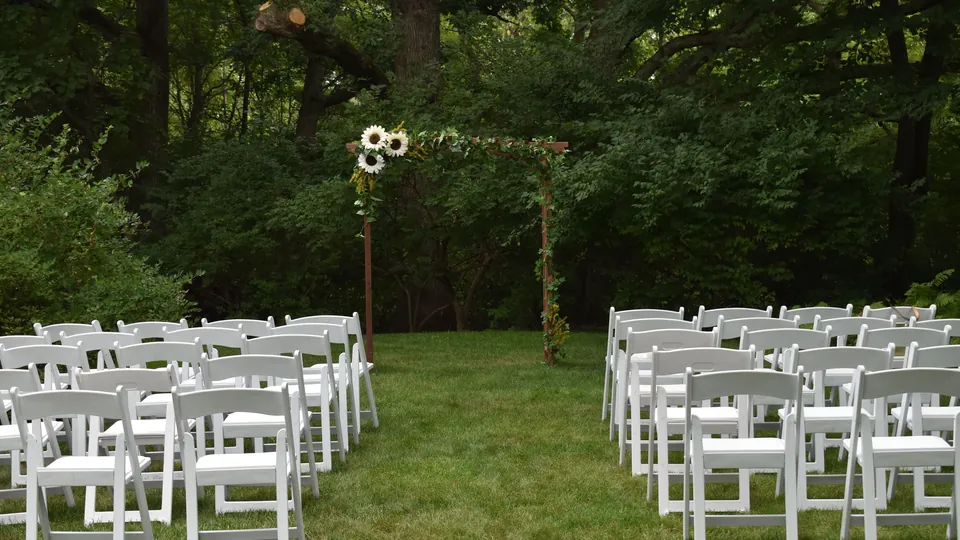 Rows of white chairs facing floral decorations in a leafy outdoor wedding venue.