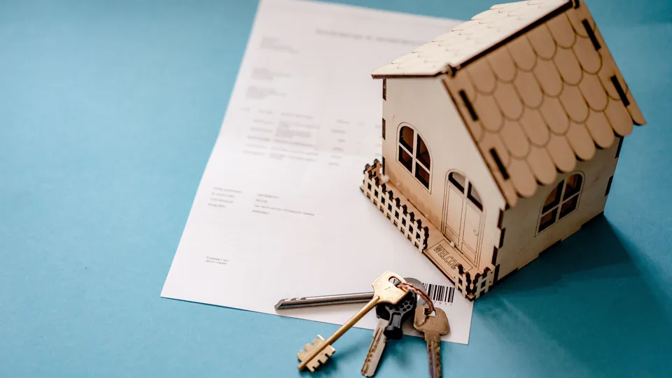 A small wooden model house and keys resting on paperwork.