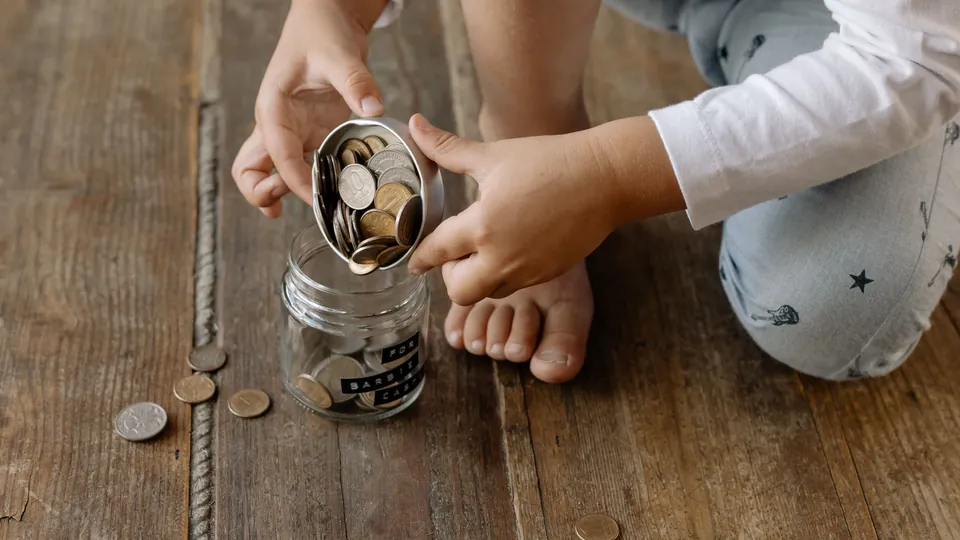 A child placing coins into a glass savings jar on a wooden floor.