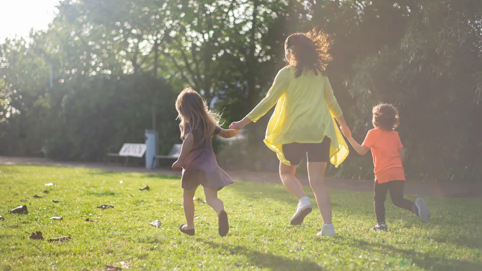 Mother holding hands with her two children as they run in a sunny park.