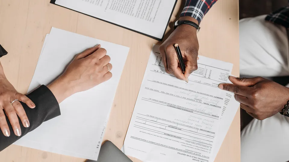 Two people reviewing and signing documents at a wooden desk.